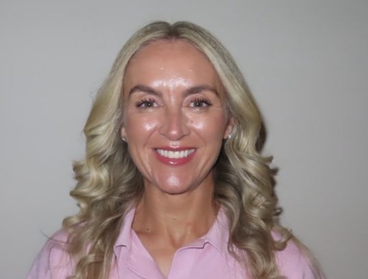 The picture is of Sarah on a white background. She has blonde hair to her shoulders and is wearing a pink top. She is smiling.